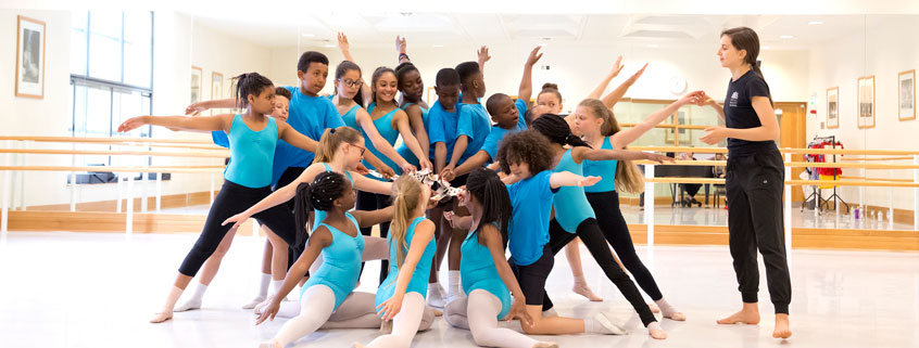 Primary Steps Large group of young, Primary Steps students in a circle, each with a hand in the centre, being watched by their teacher.